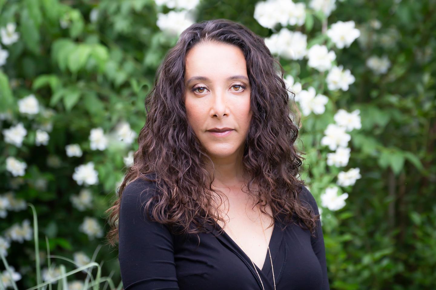 A photo of a woman with brown curly, shoulder-length hair with white flowers behind her.