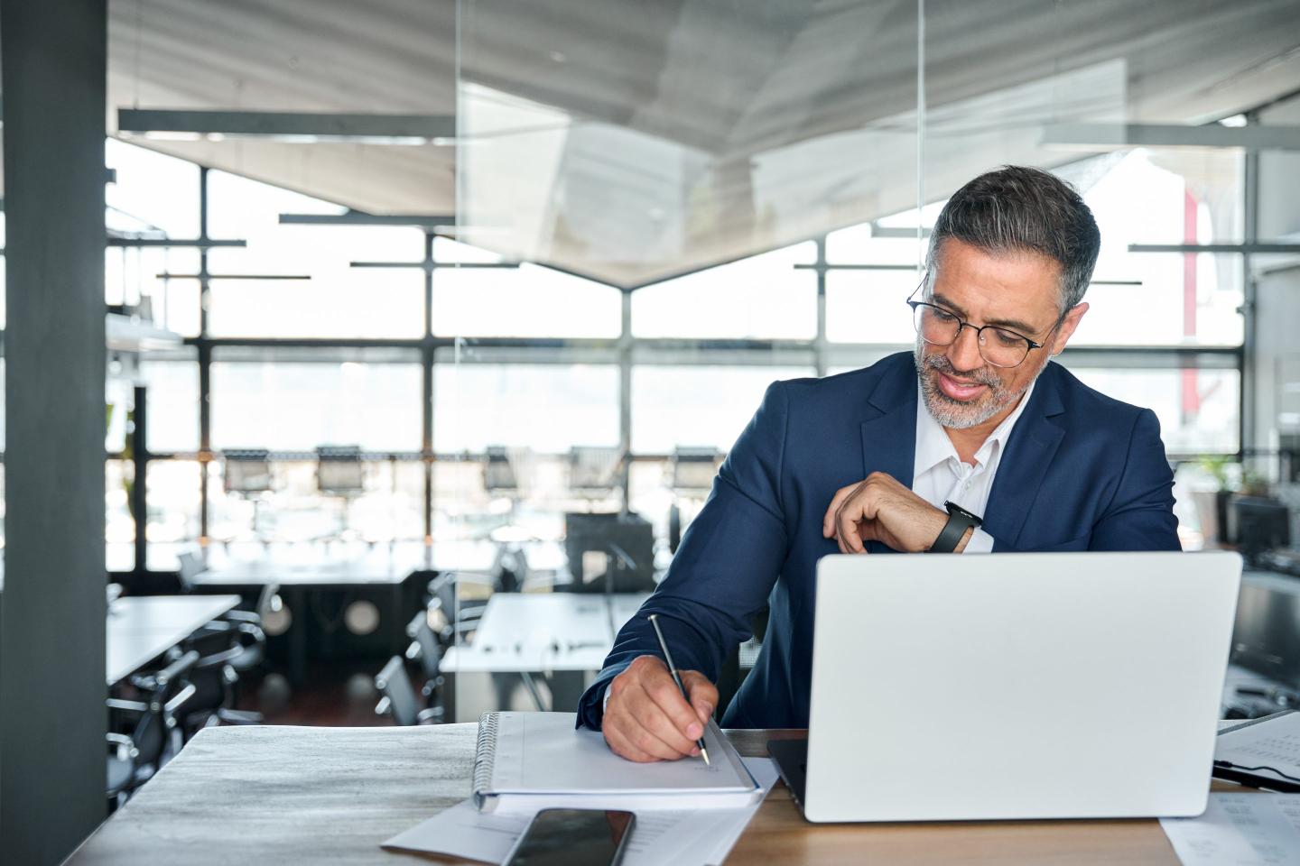 Business man takes notes on a notepad from information on his laptop.