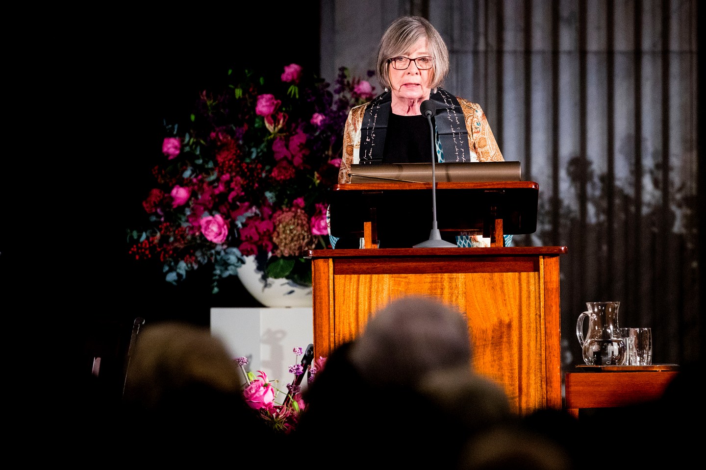 An older woman with gray hair speaks behind a wood podium in front of an audience
