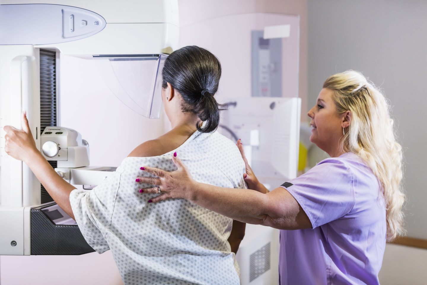 A mature African-American woman in her 40s wearing a hospital gown, getting her annual mammogram. She is being helped by a technologist, a blond woman wearing scrubs.