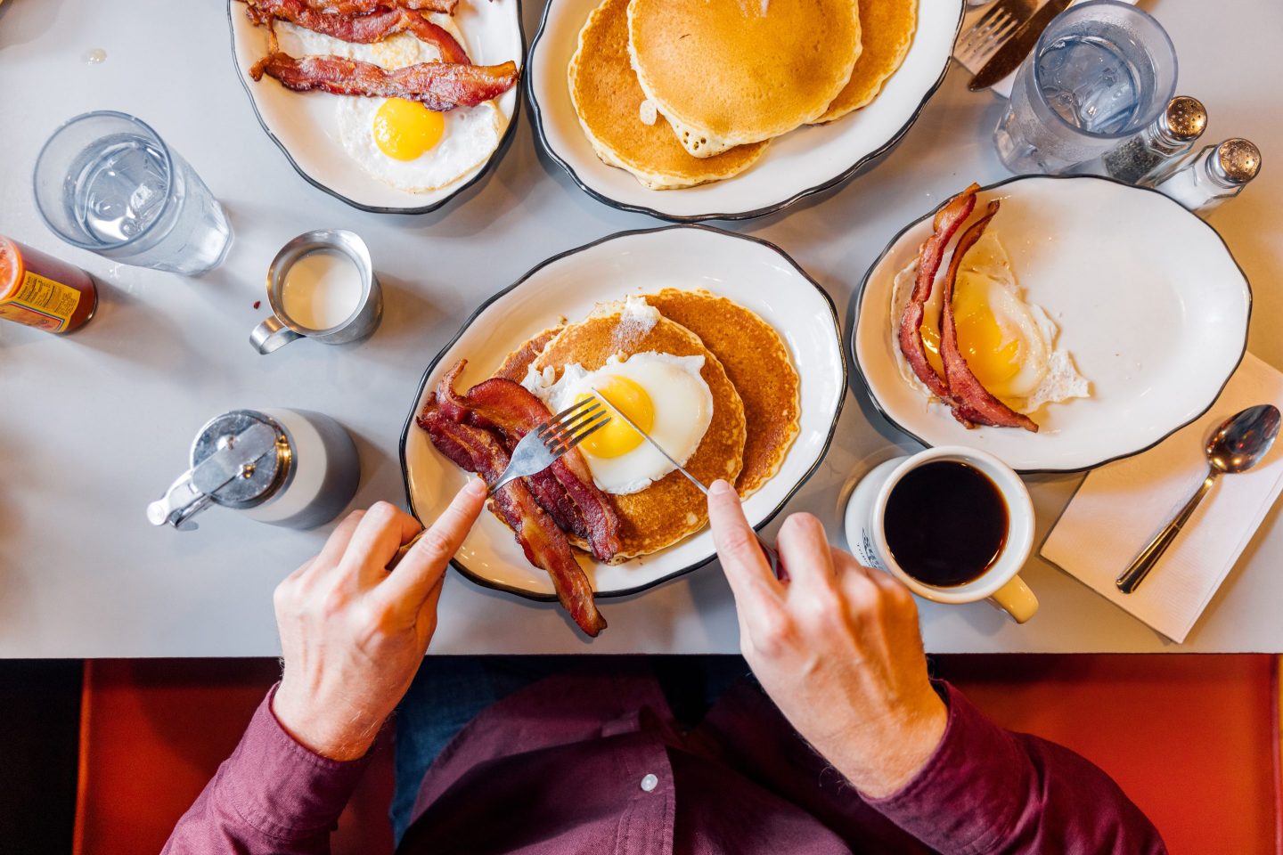 An unseen person eats a plate of pancakes, fried eggs and bacon in a traditional American diner. A cup of black coffee is seen beside his food.