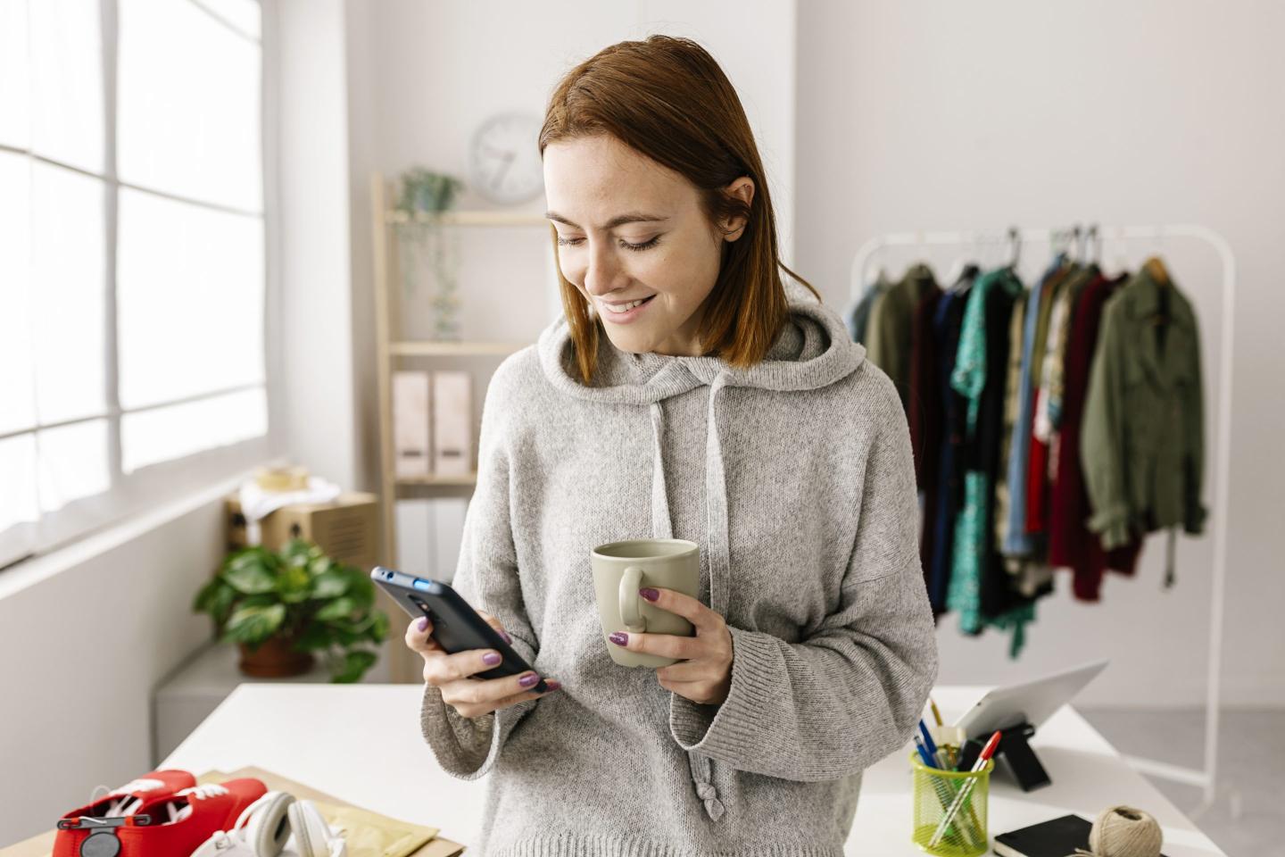 Female owner using cell phone and drinking coffee while standing in home office