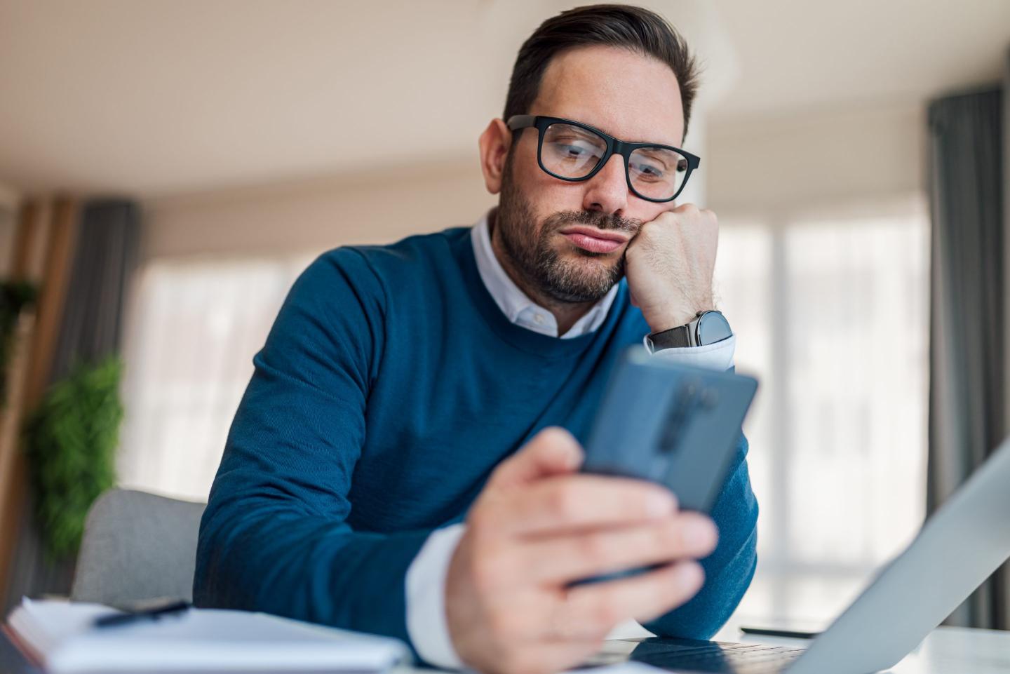 A man in a blue sweater looks at his smartphone while working