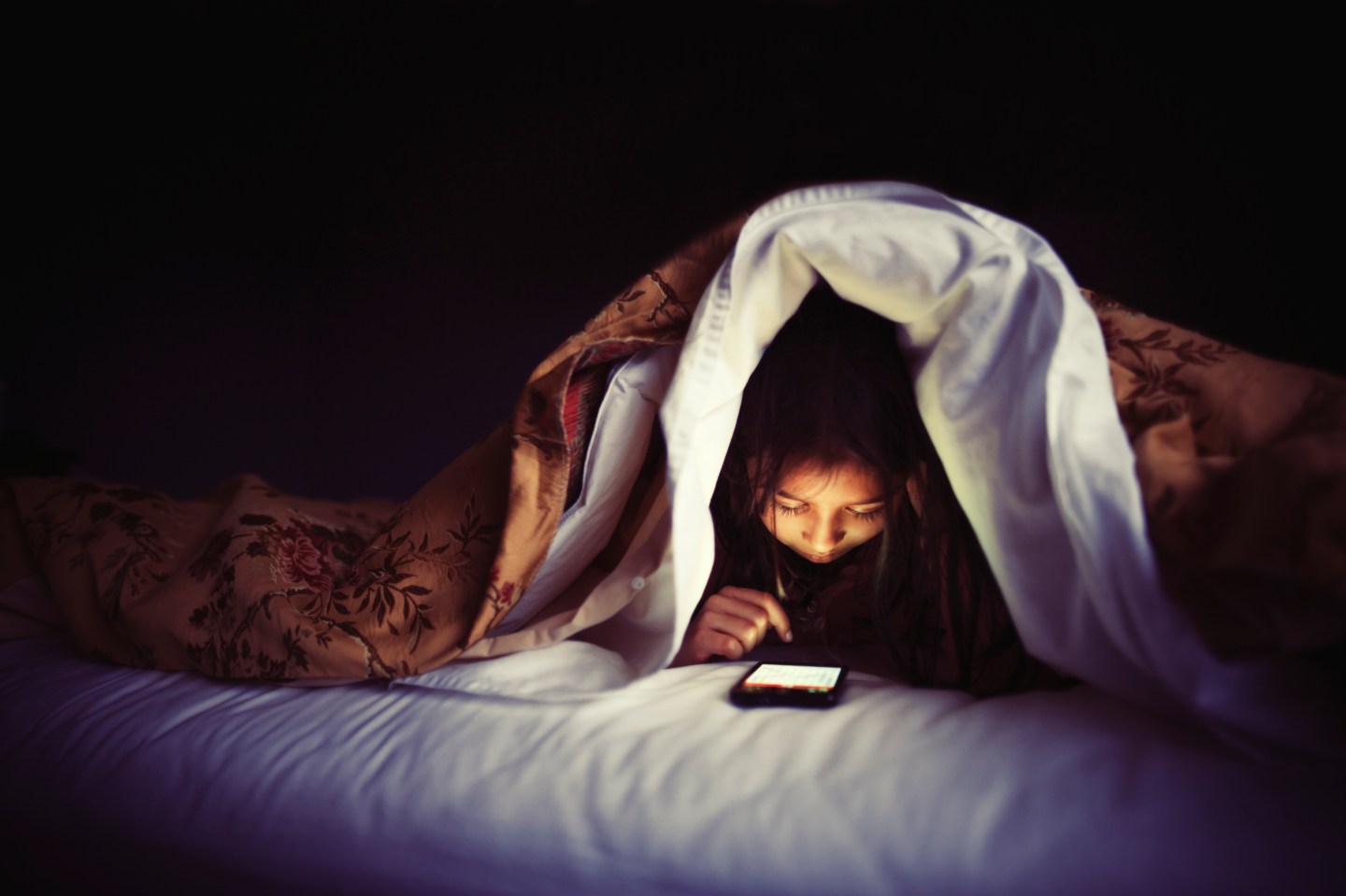 Girl using smartphone under bed covers.