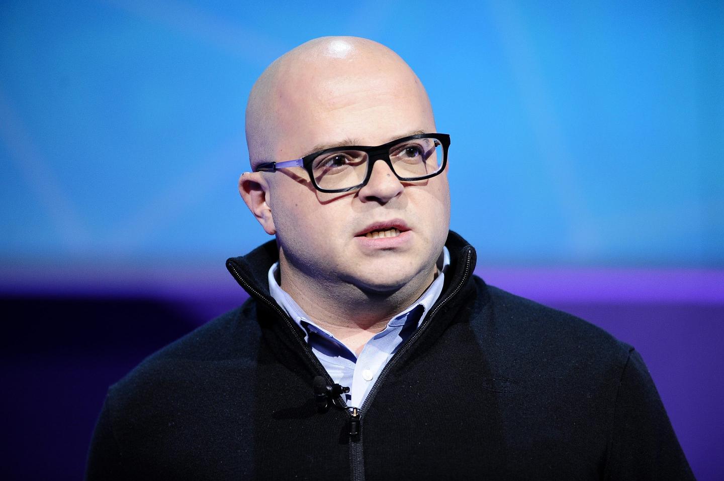 Jeff Lawson, CEO of Twilio, is standing in front of a blue and purple screen with black glasses, blue button up underneath a black pullover.