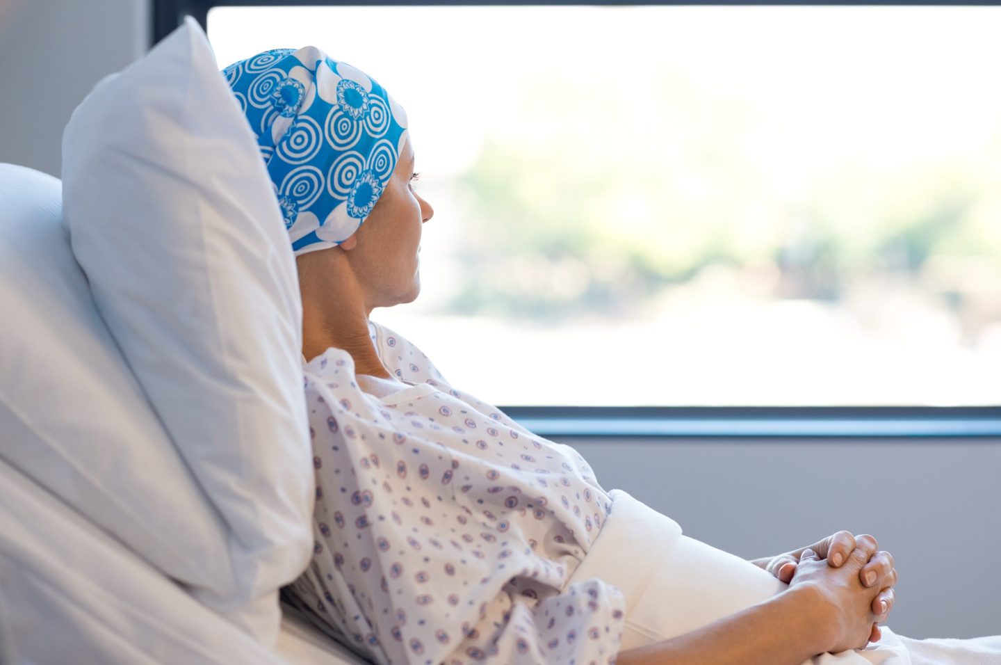 a woman in a hospital bed looks out a window. she wears a blue headscarf