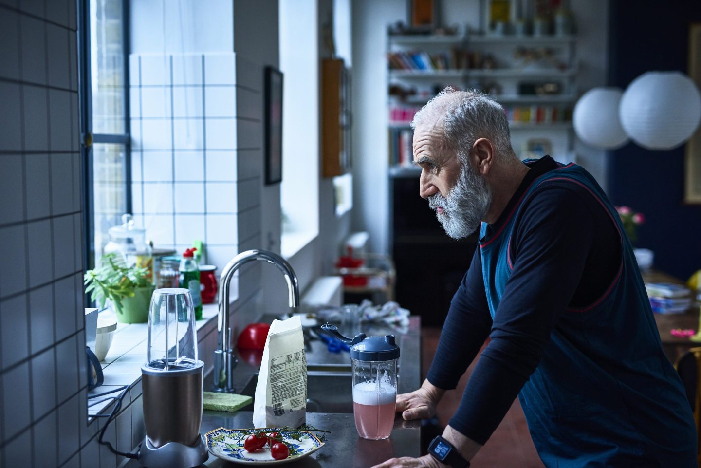 Senior man standing over a kitchen sink looking out a window with a defeated look on his face