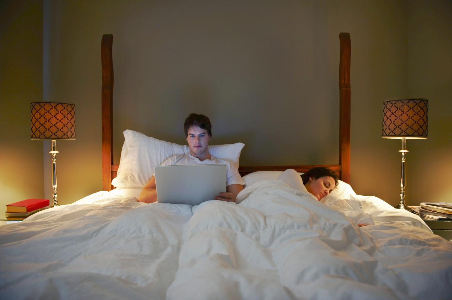 Man in bed on laptop while woman is sleeping.