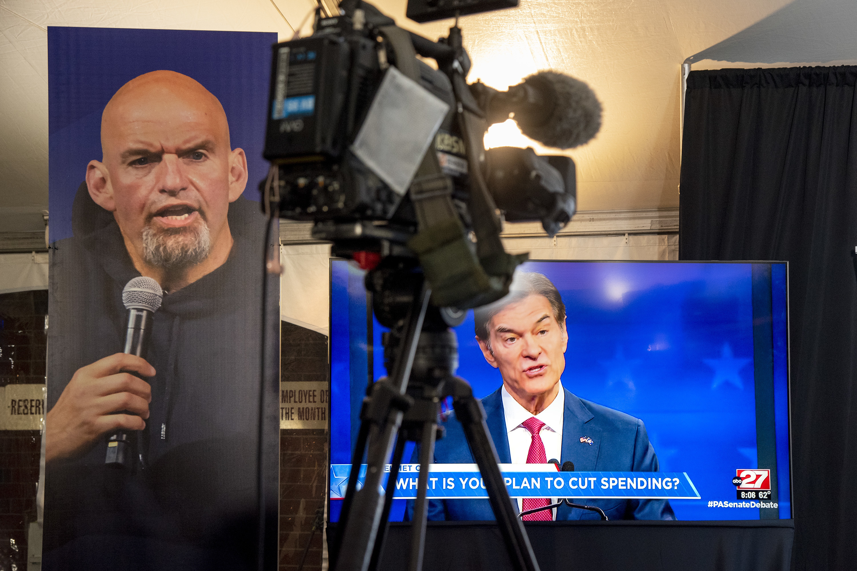 Republican Mehmet Oz, right, is seen live on a monitor in the media tent, next to a poster of Democrat John Fetterman, left, as the two U.S. Senate candidates hold their first and only debate, at the WHTM-TV/ABC 27 Studio in Harrisburg, Pa., Oct. 25, 2022.