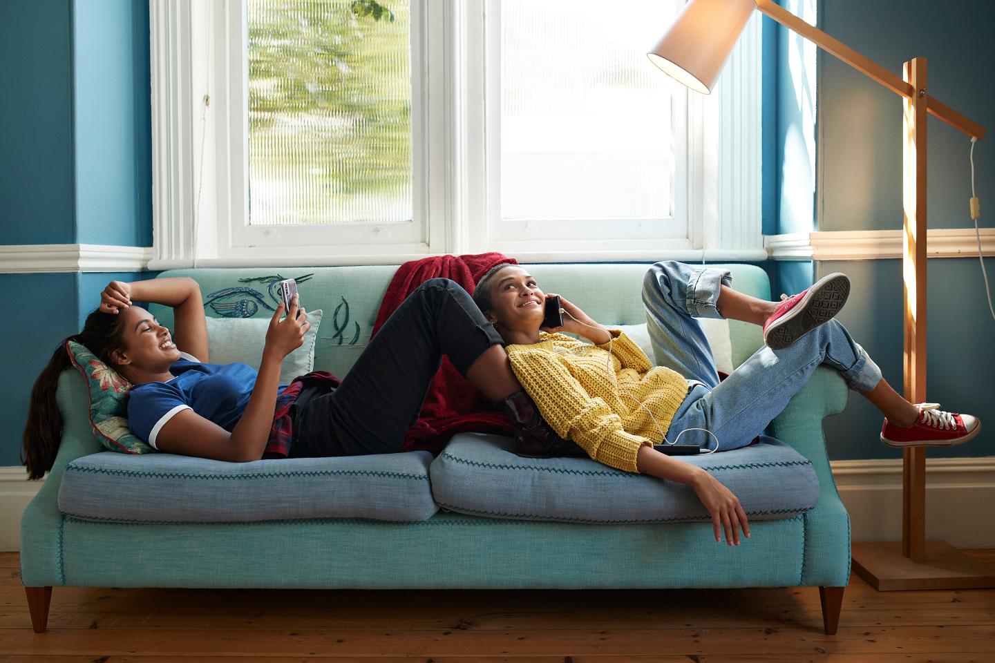 Full length of smiling friends using phones while resting together on sofa at home
