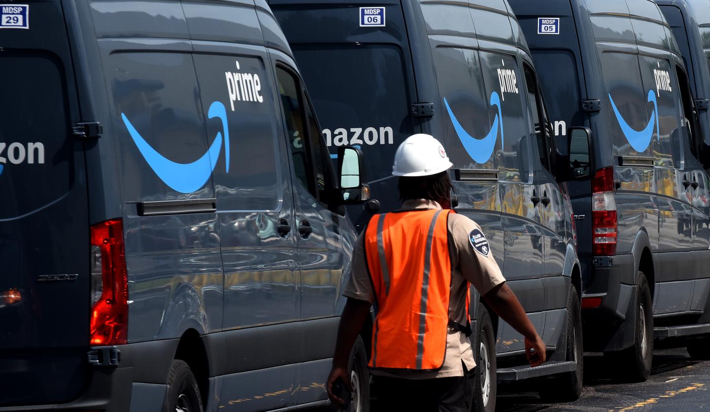 Amazon worker in a bright orange vest walking away from the camera and past a line of Amazon vans.