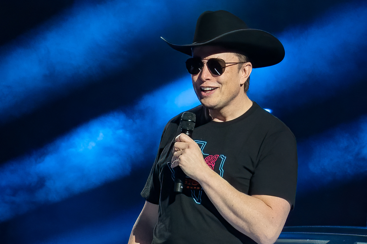 Twitter CEO Elon Musk wearing a cowboy hat, sunglasses, and dark tshirt with a mic in hand.