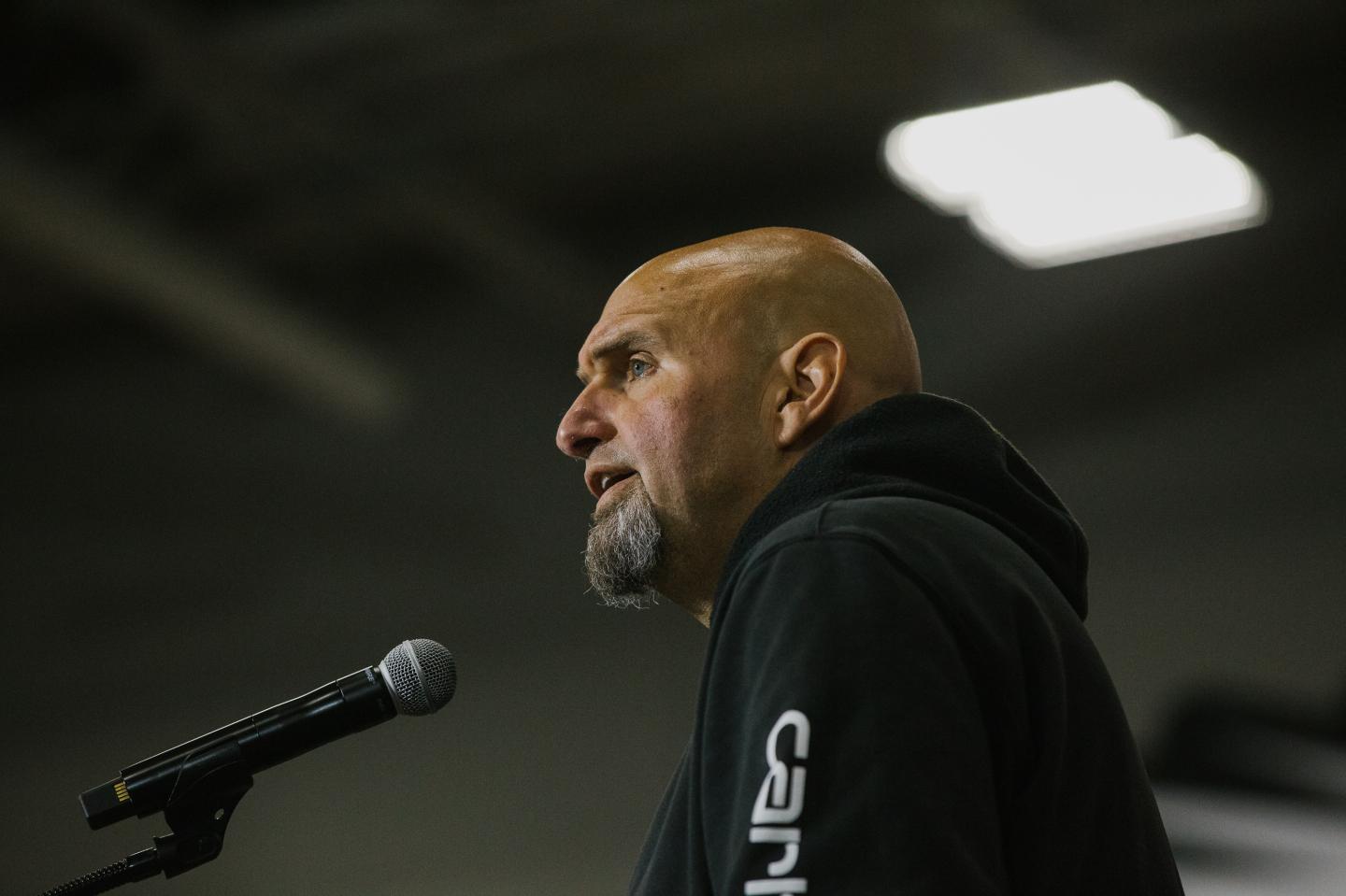 John Fetterman speaking in front of a microphone while standing at a podium wearing a black hooded sweathshirt.