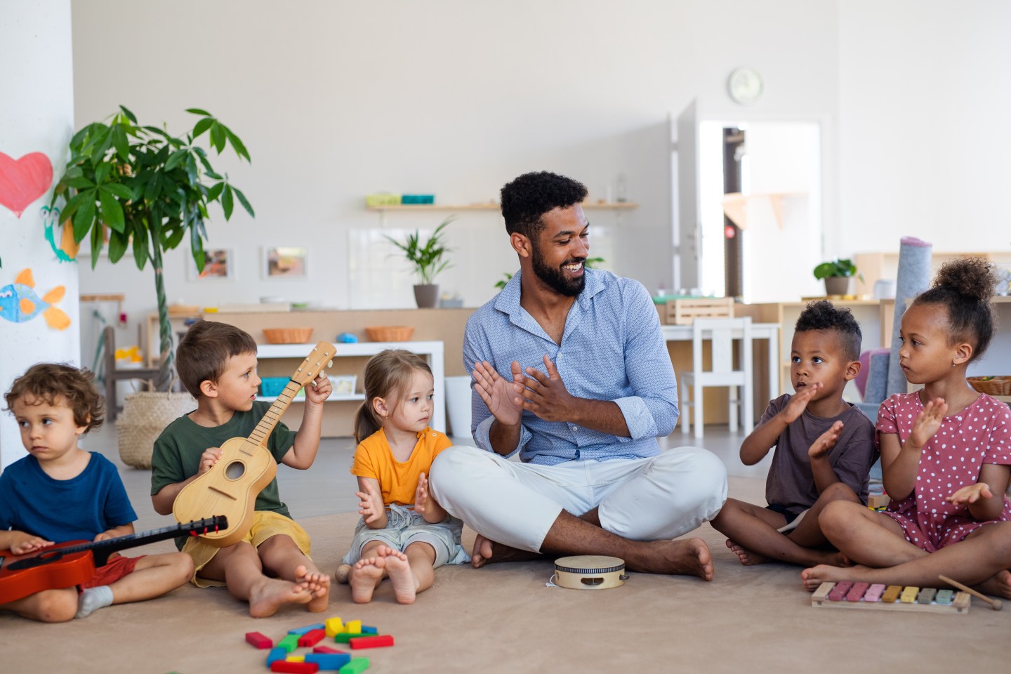 Group of small nursery school children with man teacher sitting on floor indoors in classroom, playing musical instruments.