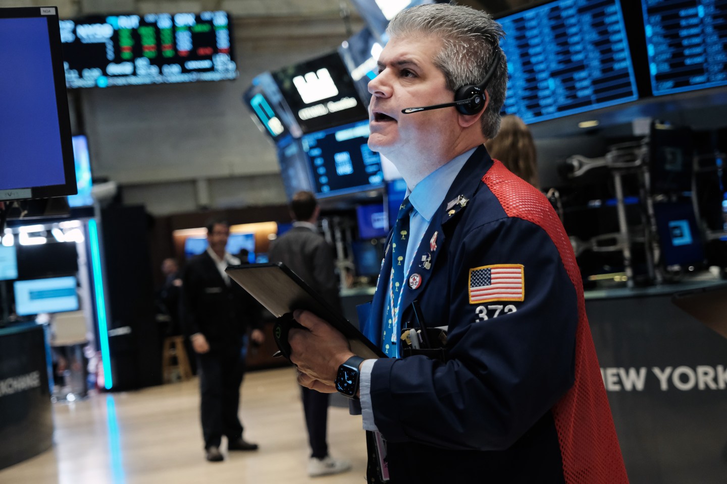 A trader on the floor of the New York Stock Exchange looking up at a screen and speaking into an earpiece.