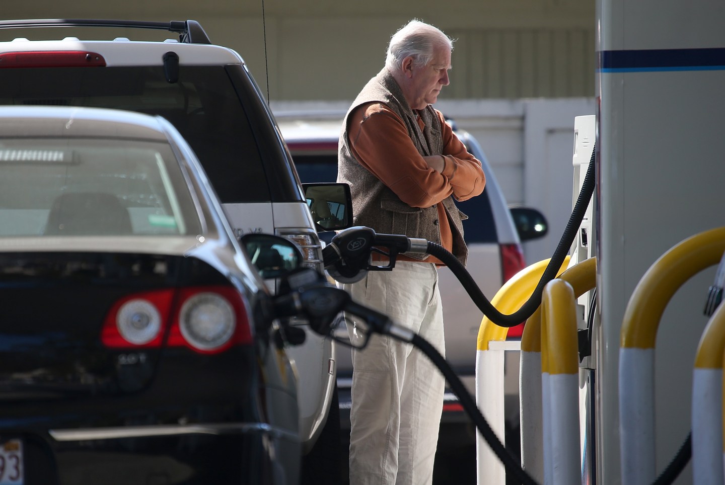 A senior man pumps gasoline into his car at a gas station with a concerned look on his face