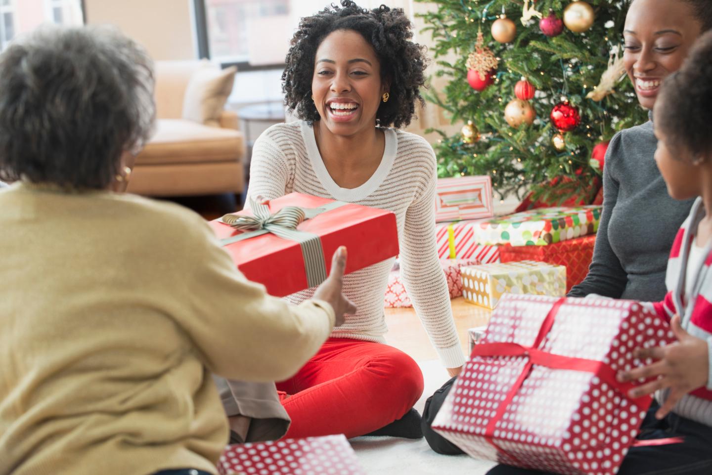 A young woman is seen giving a holiday gift to an older relative as they sit in front of a Christmas tree with their family.
