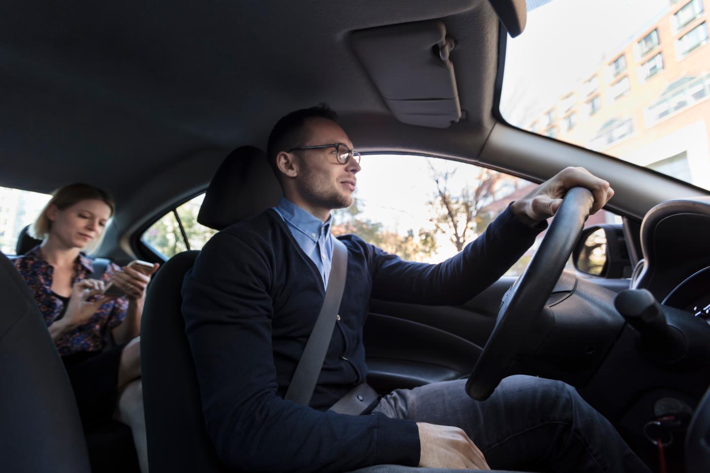 A young man driving a car with a person in the backseat.