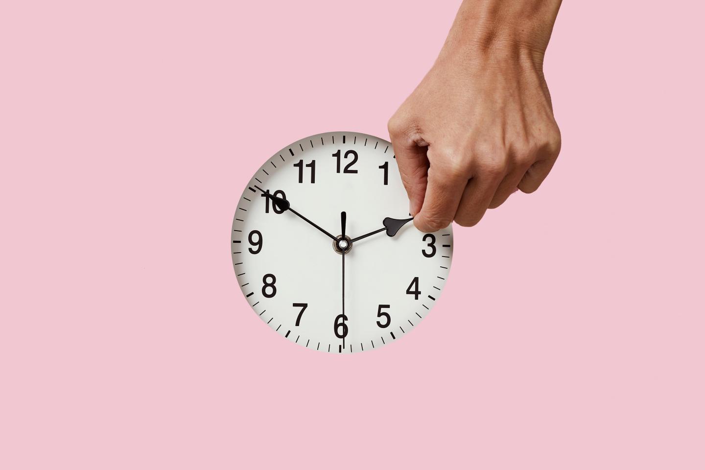 closeup of the hand of a young caucasian man setting a clock backward or forward, at the end or at the beginning of the summer, on a pink background