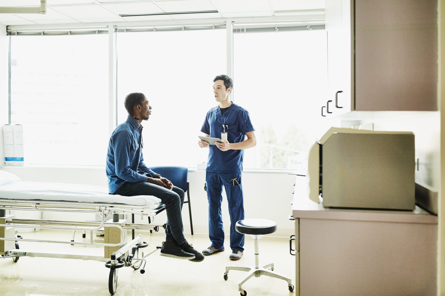 Male nurse holding digital tablet consulting with patient in exam room.