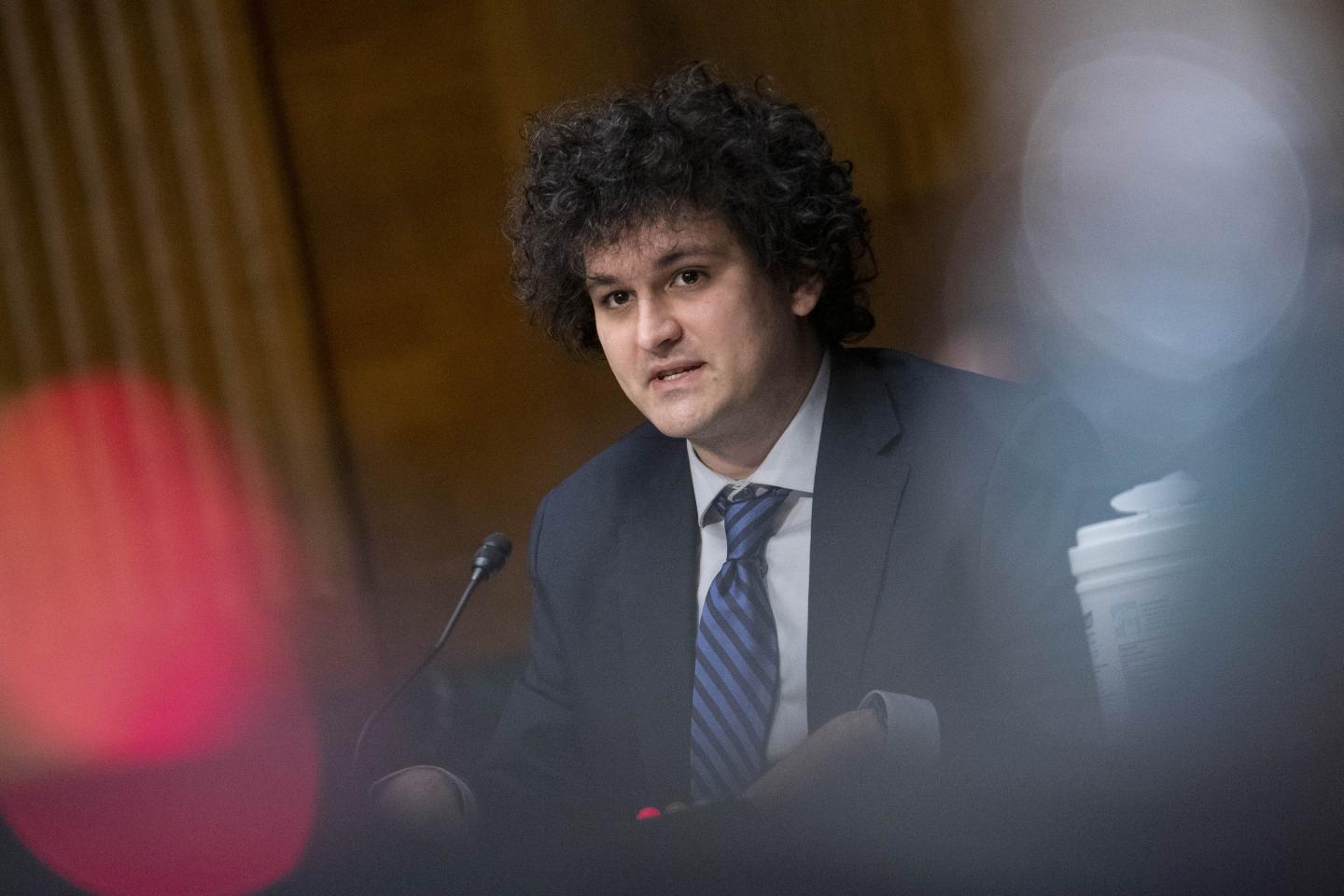 A white man with black curly hair and wearing a dark suit with a blue tie speaks into a microphone