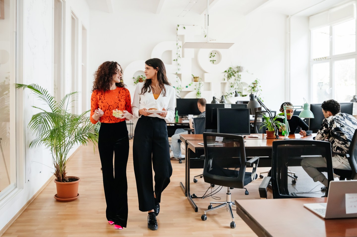 Two women, one in a red blouse and the other in a white one, walk past desks in a spacious office with tall ceilings