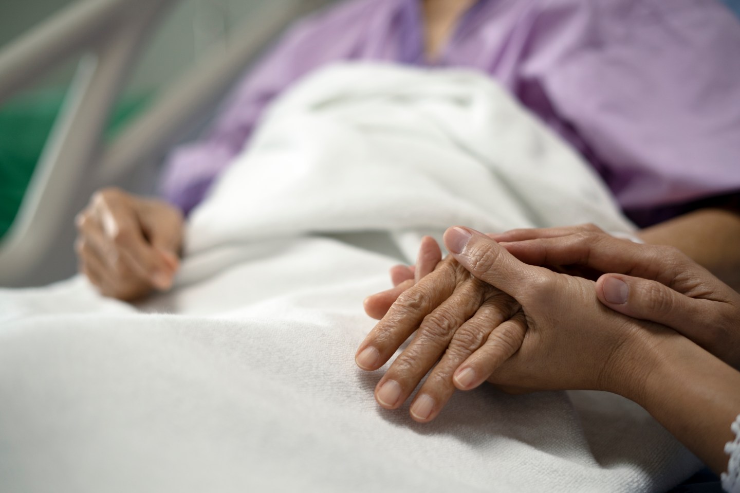 A set of hands holding the hand of a person laying in a hospital bed.