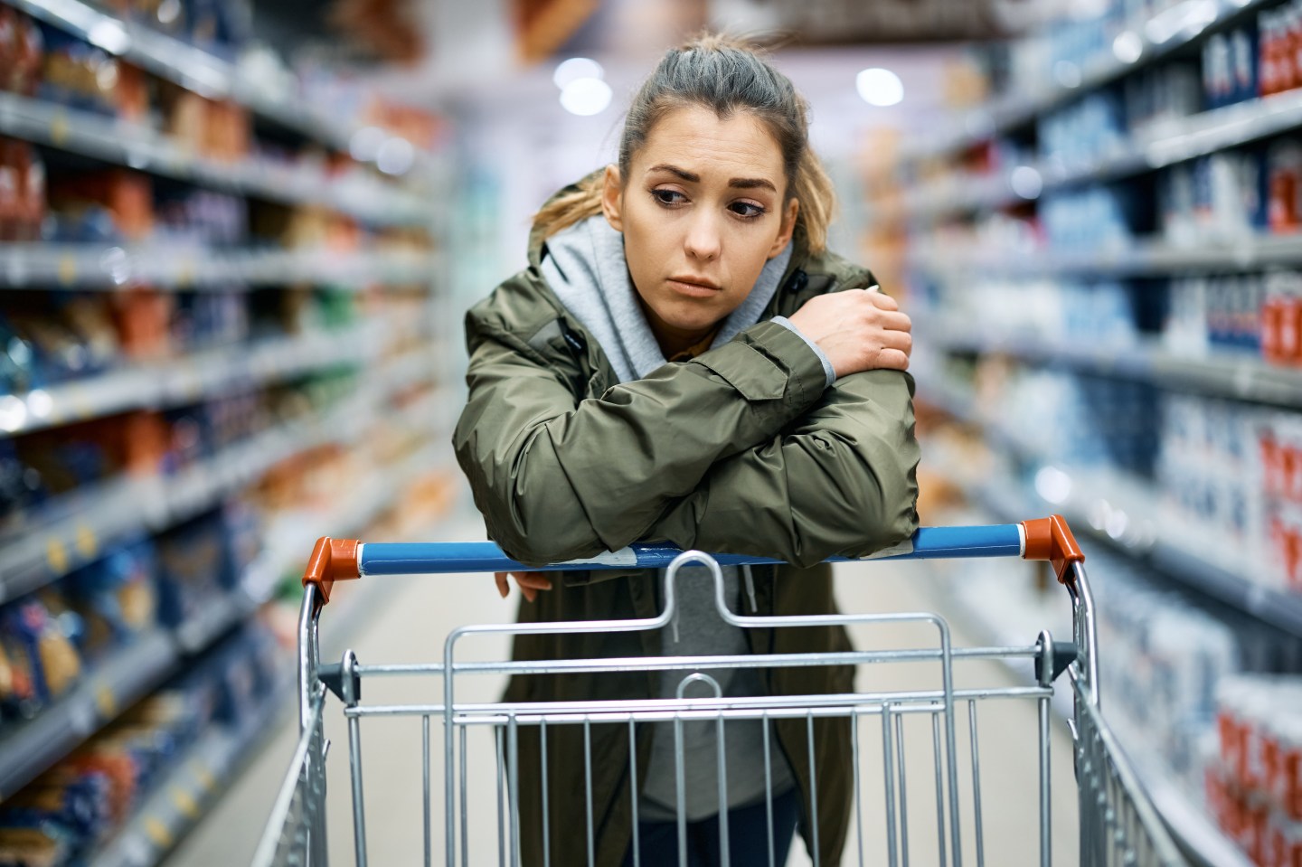 A woman looking sad while leaning on a grocery cart in a supermarket aisle.