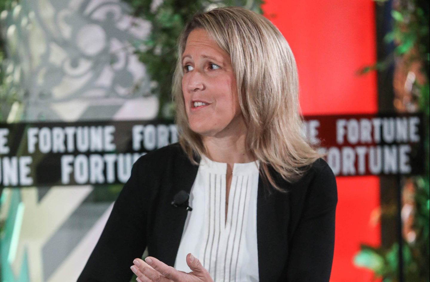 A blonde woman in a white blouse and black cardigan speaks to someone out of view.