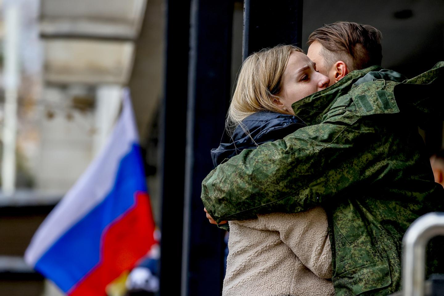 A Russian man dressed in a military uniform embracing a woman in front of a Russian flag