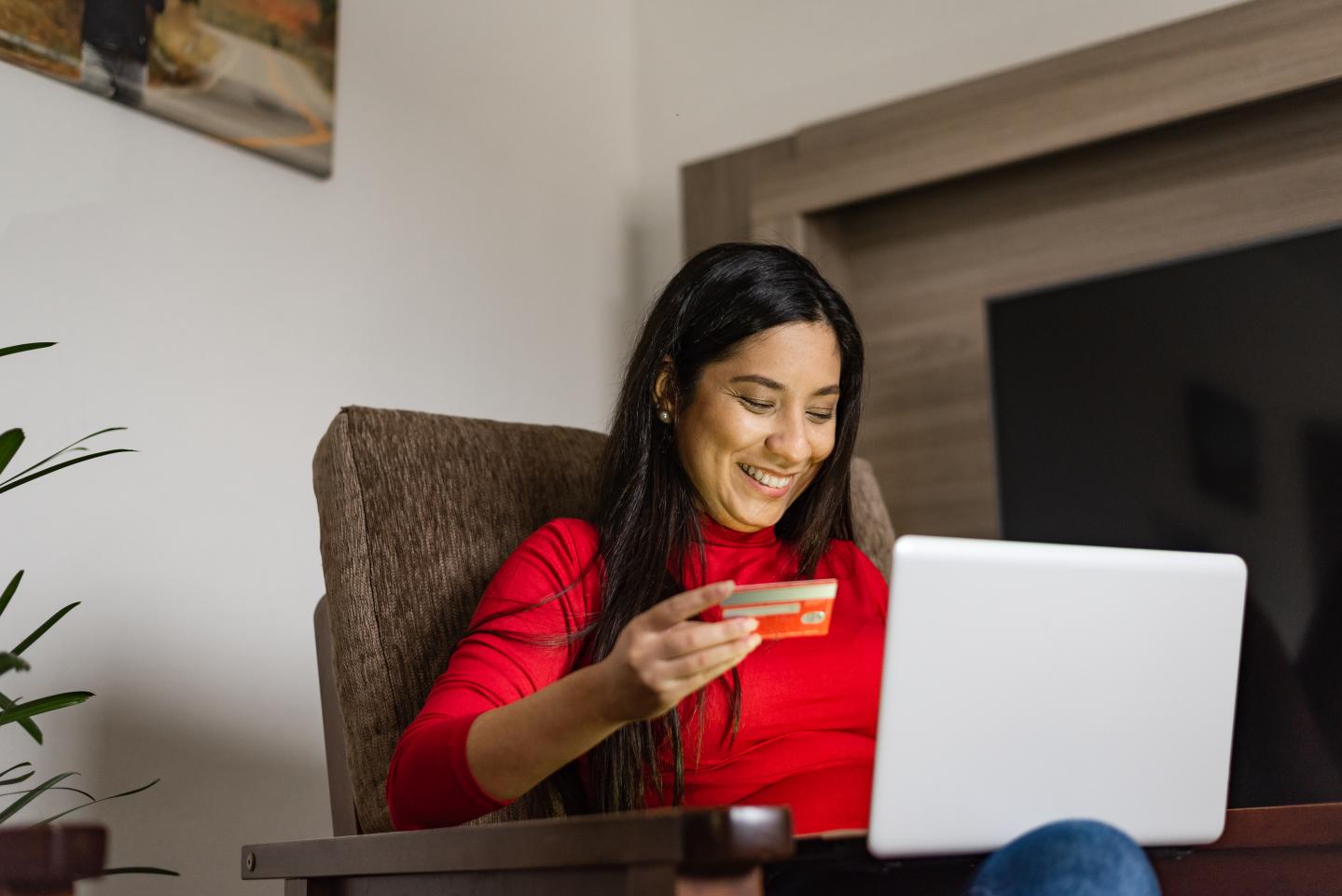 Young woman sitting on the armchair holding a credit card for online shopping on laptop