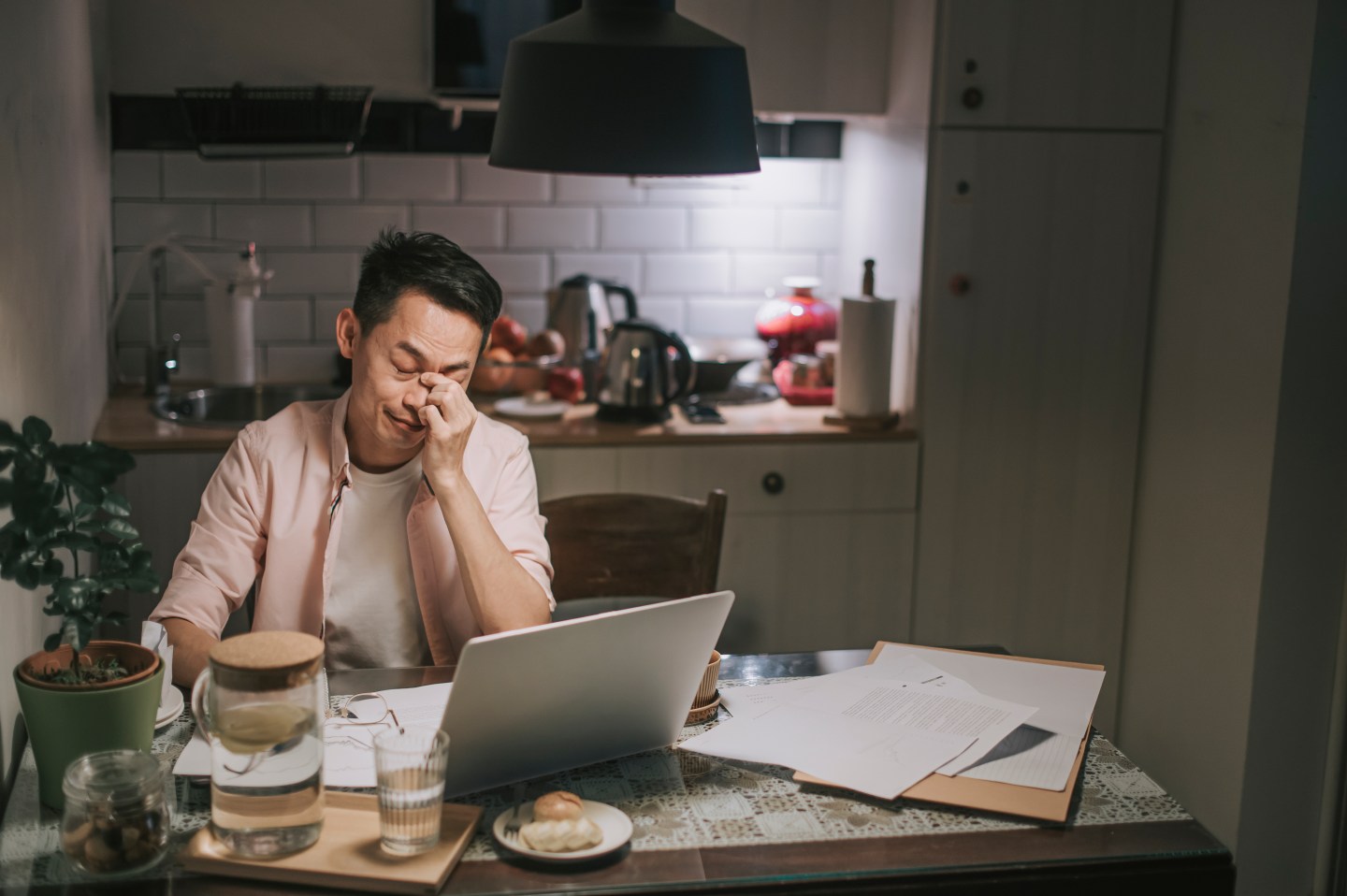 Distressed man working on a laptop on a kitchen table covered in papers