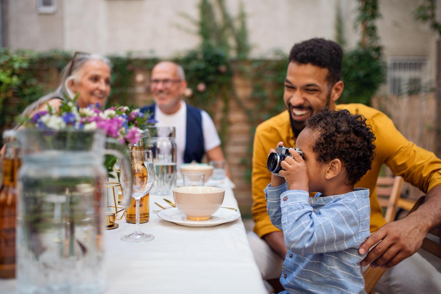 Little multiracial boy with camera sitting and taking picture outdoors in garden during birthday party.