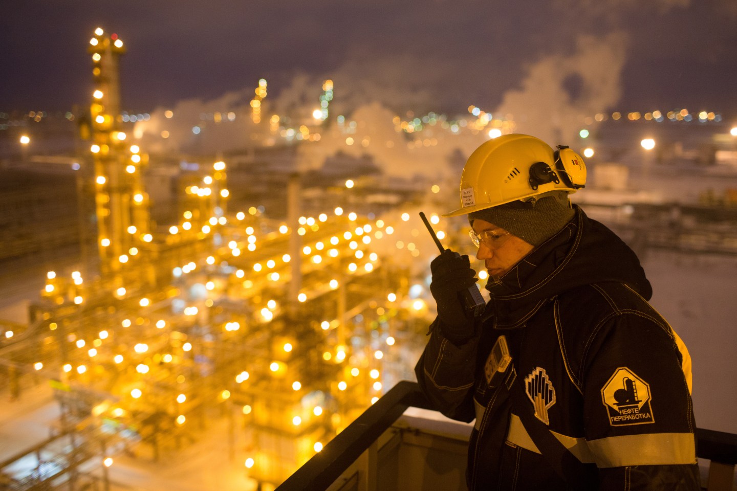 An engineer talking into a receiver at night overlooking an illuminated oil refinery plant in Russia