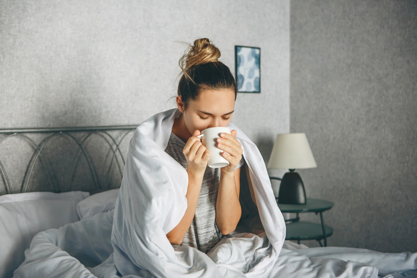 Young woman drinks from a mug in bed wrapped in a blanket.