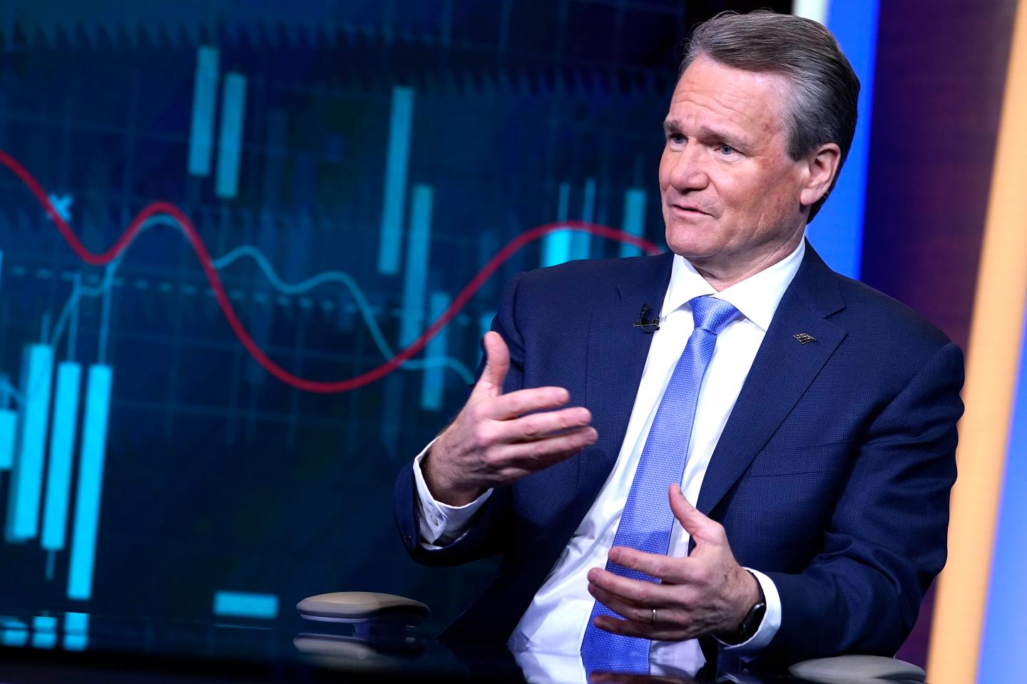 A white man dressed in a blue suit and tie is talking to someone off-camera with his hands and a chart of stock price increases and decreases behind him on a screen