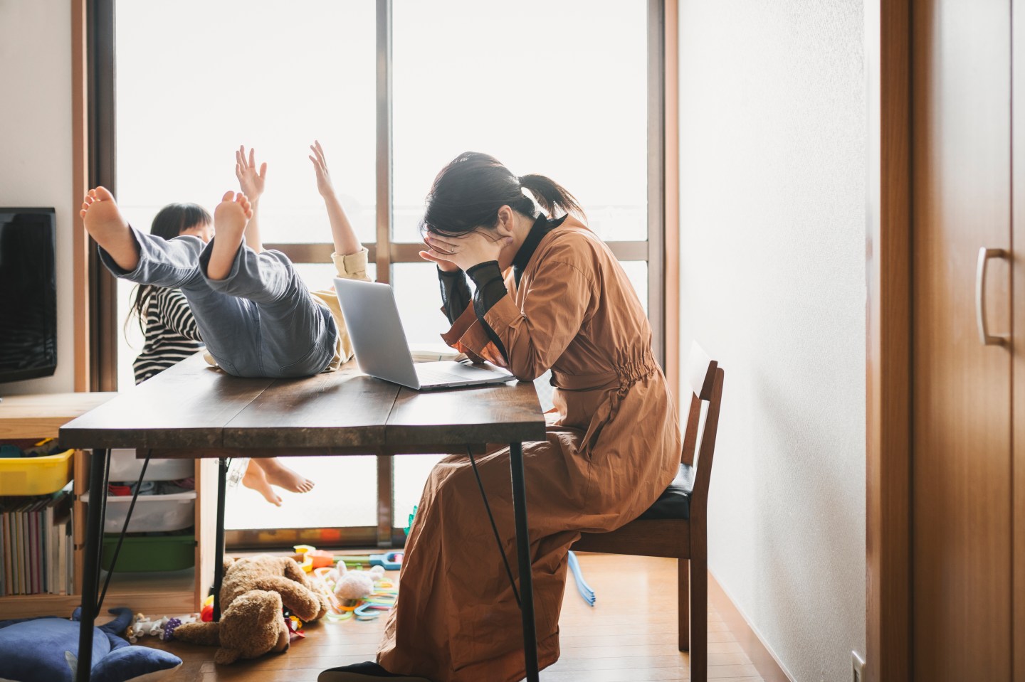 Mother working from home on her laptop holding her head in her hands while her kids play around her