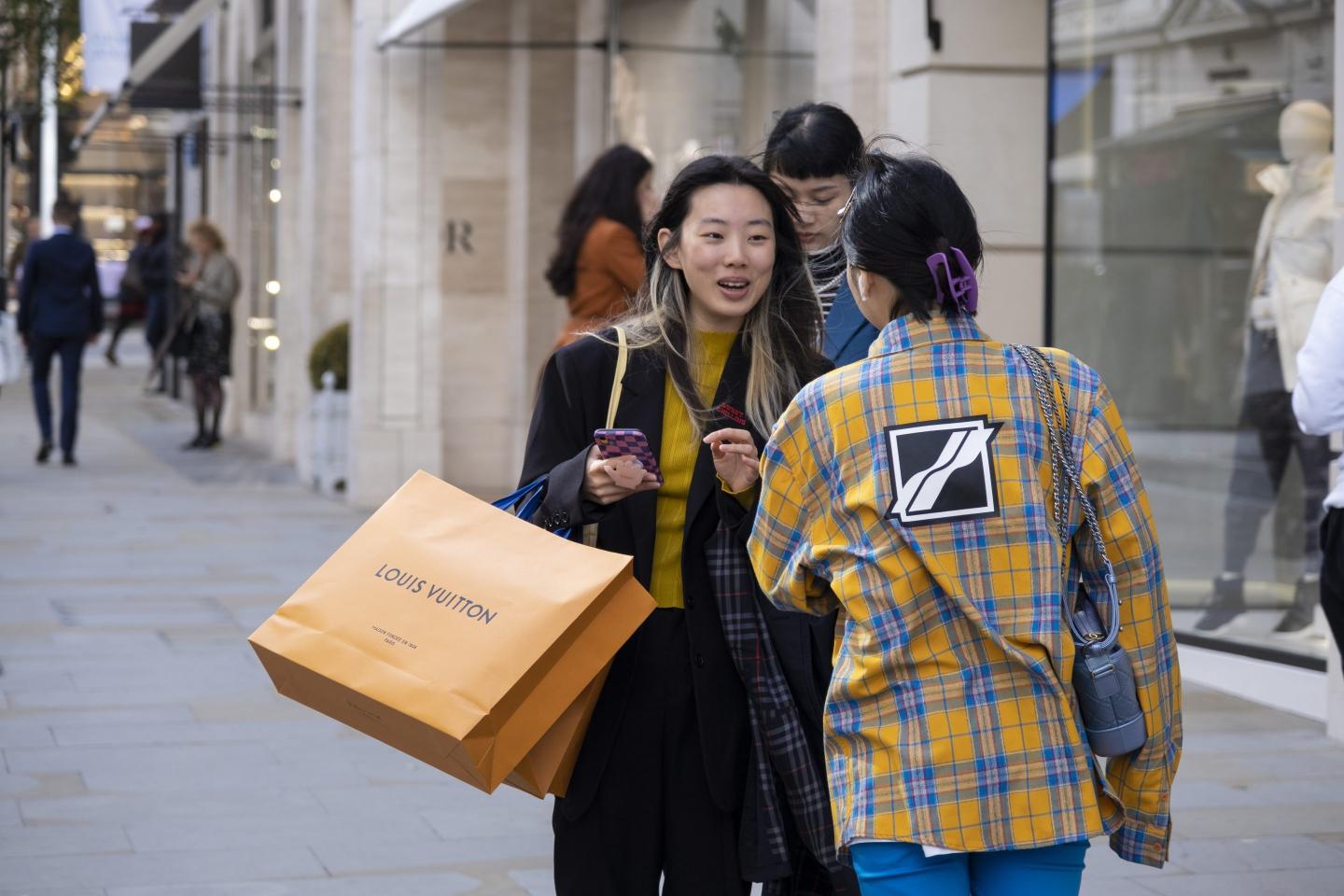 Two people - one with a large Louis Vuitton shopping bag - stand speaking in the street.