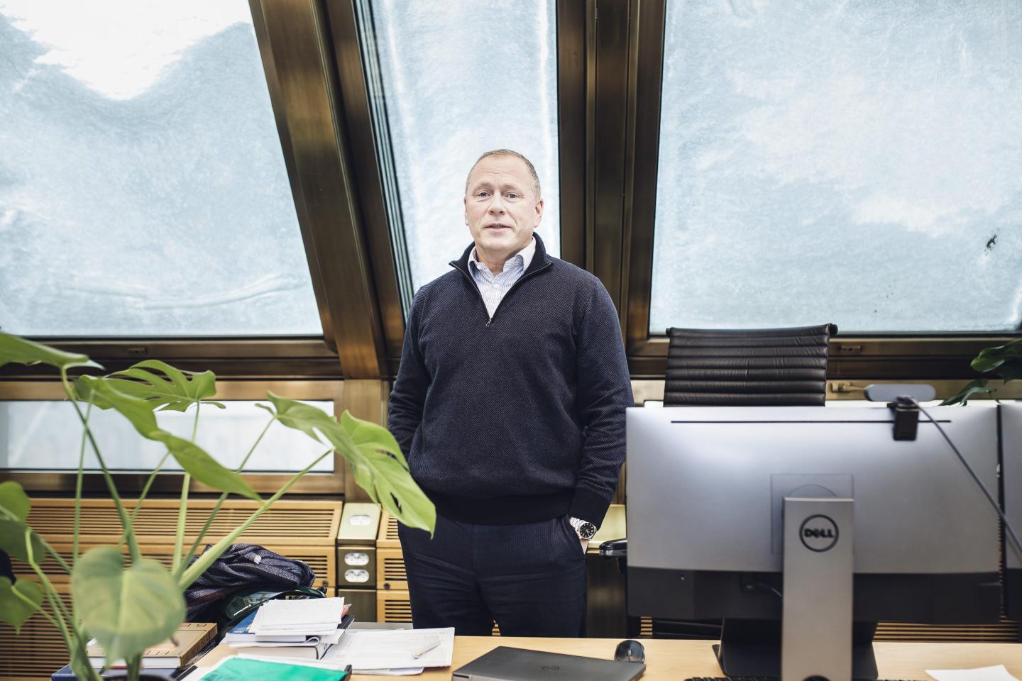 A white man in a blue collared shirt and a pullover stands with his hands in his pockets behind a desk with a plant and computer on it