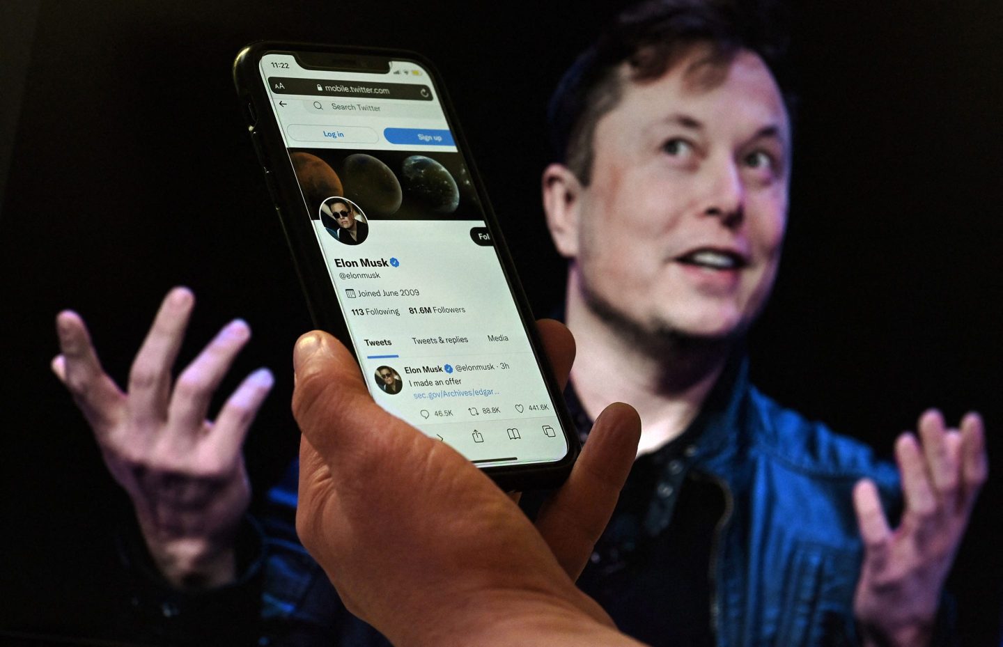 a phone screen displays the Twitter account of Elon Musk with a photo of him shown in the background