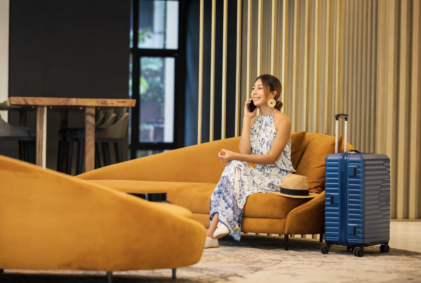 A woman sits on a sofa on the phone, a suitcase beside her.