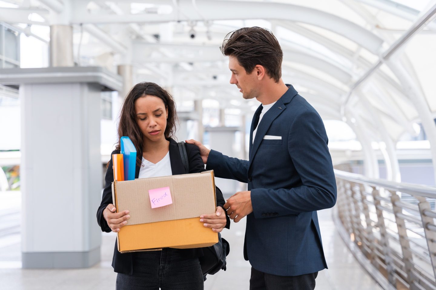 A young man consoles a young woman holding a box filled with binders and personal belongings.