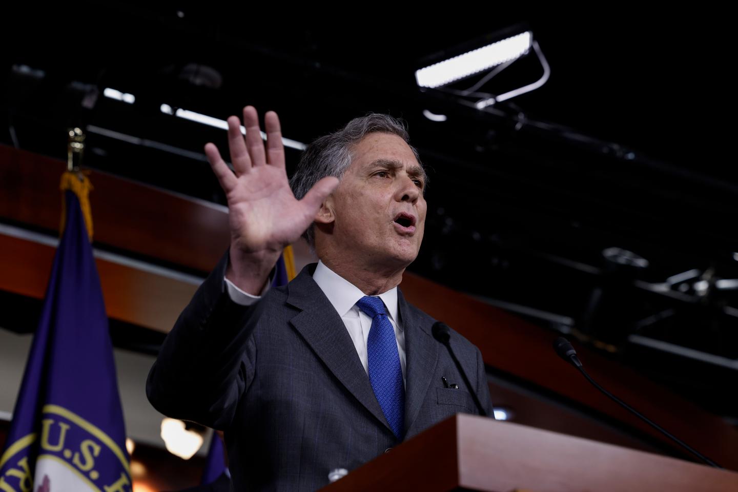 A man dressed in a dark grey suit and blue tie, holds up his right hand towards an audience of reporters, while he stands behind a podium, speaking into a microphone