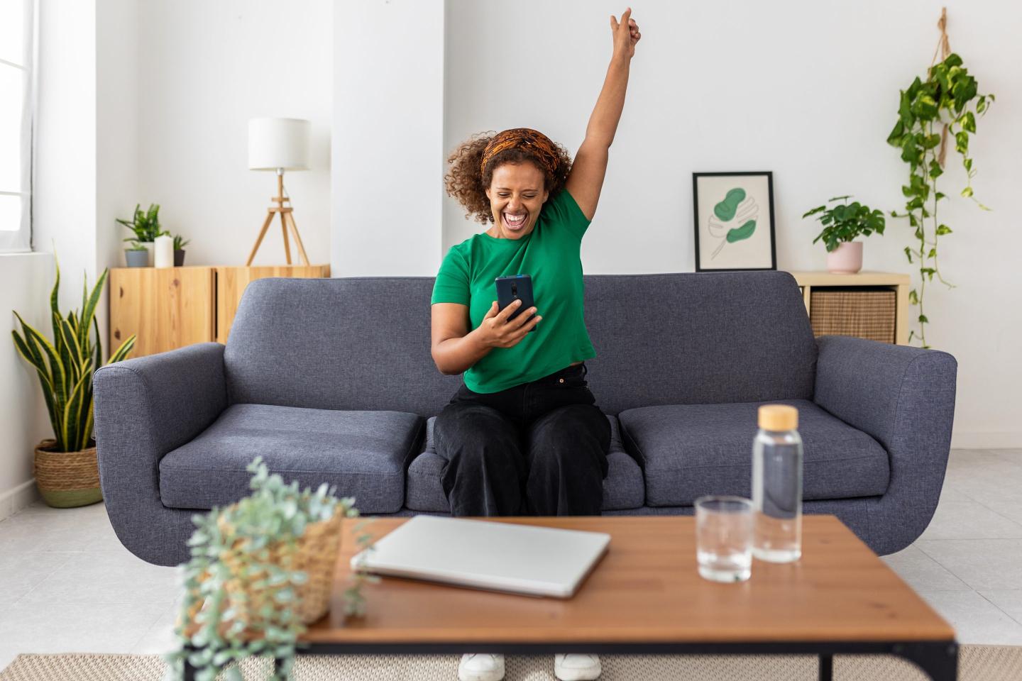 A woman sits on a sofa, cheering as she looks at her phone.
