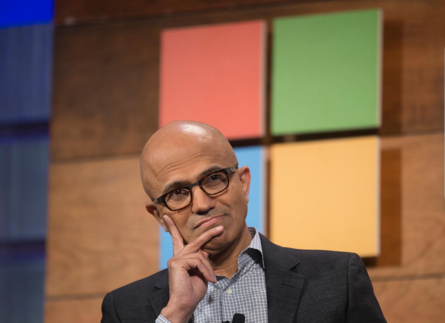 Microsoft CEO Satya Nadella on stage listening to an audience member ask a question, hand on face, sitting in front of a giant microsoft logo.
