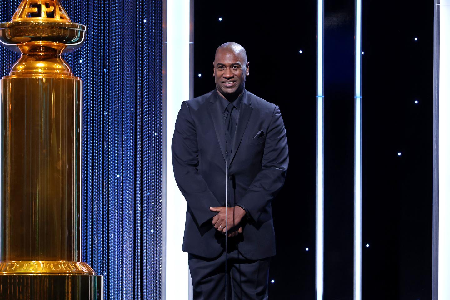 A Black man in a dark suit stands on a stage with his hands clasped before him