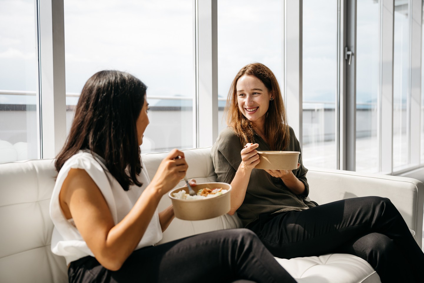 Two female co-workers are sitting on a beige sofa in a bright modern office and enjoying a healthy take-away lunch.