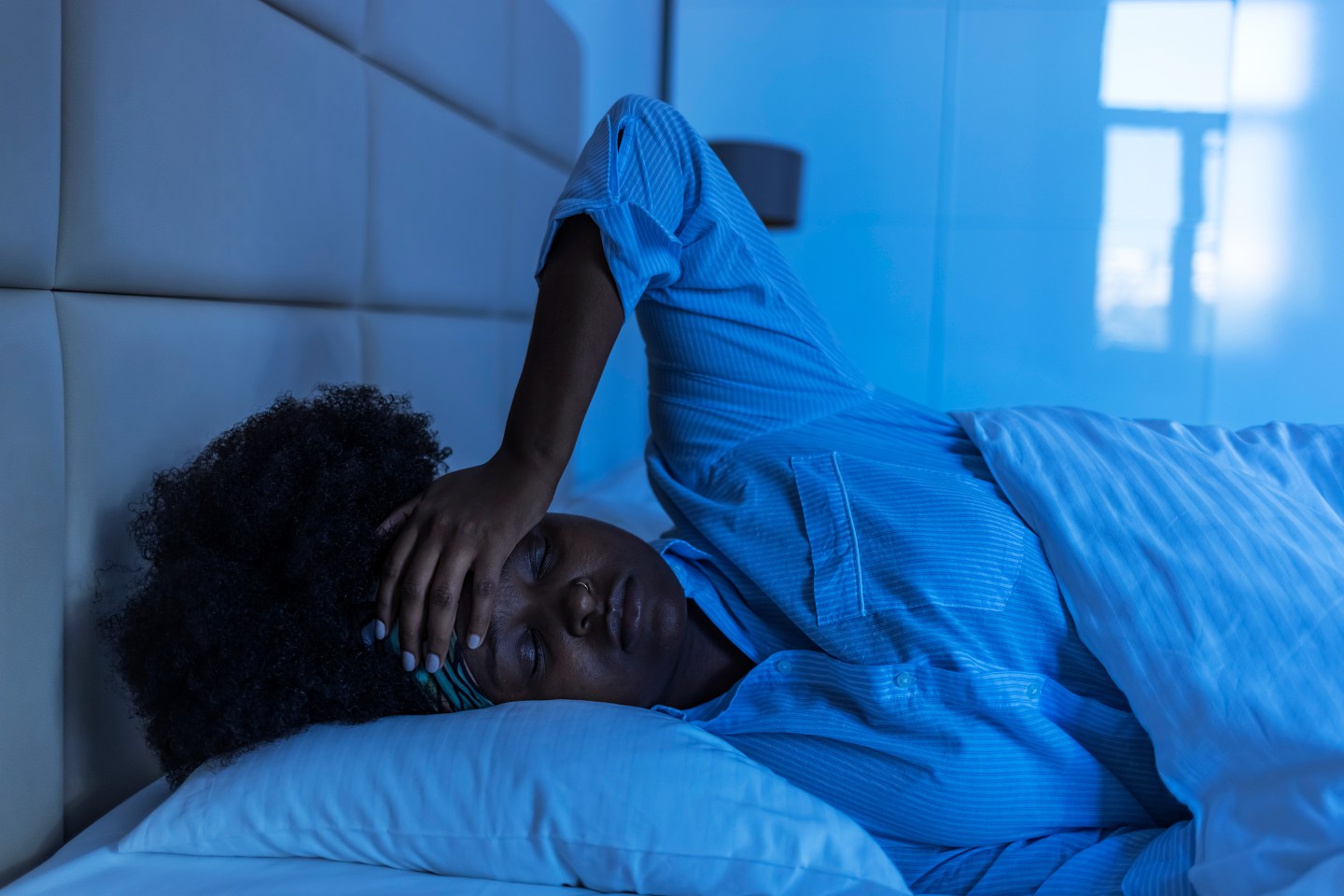 Black woman holding hand on forehead looking sad while laying in bed at night.