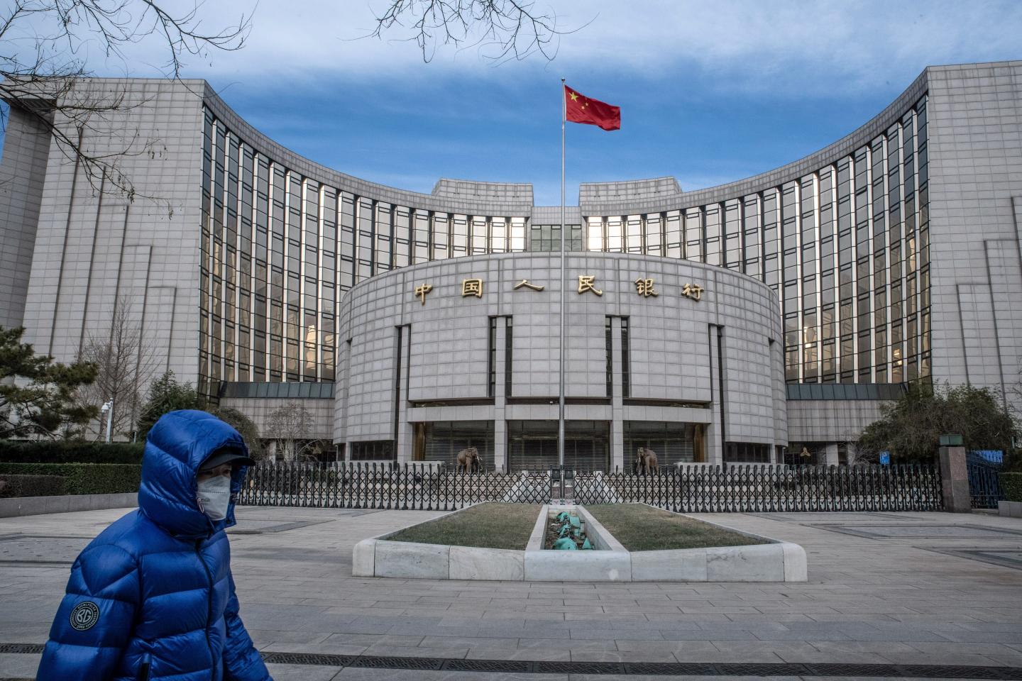 A man in a blue puffer coat walks in front of a grey concrete building with the Chinese flag blowing in the wind