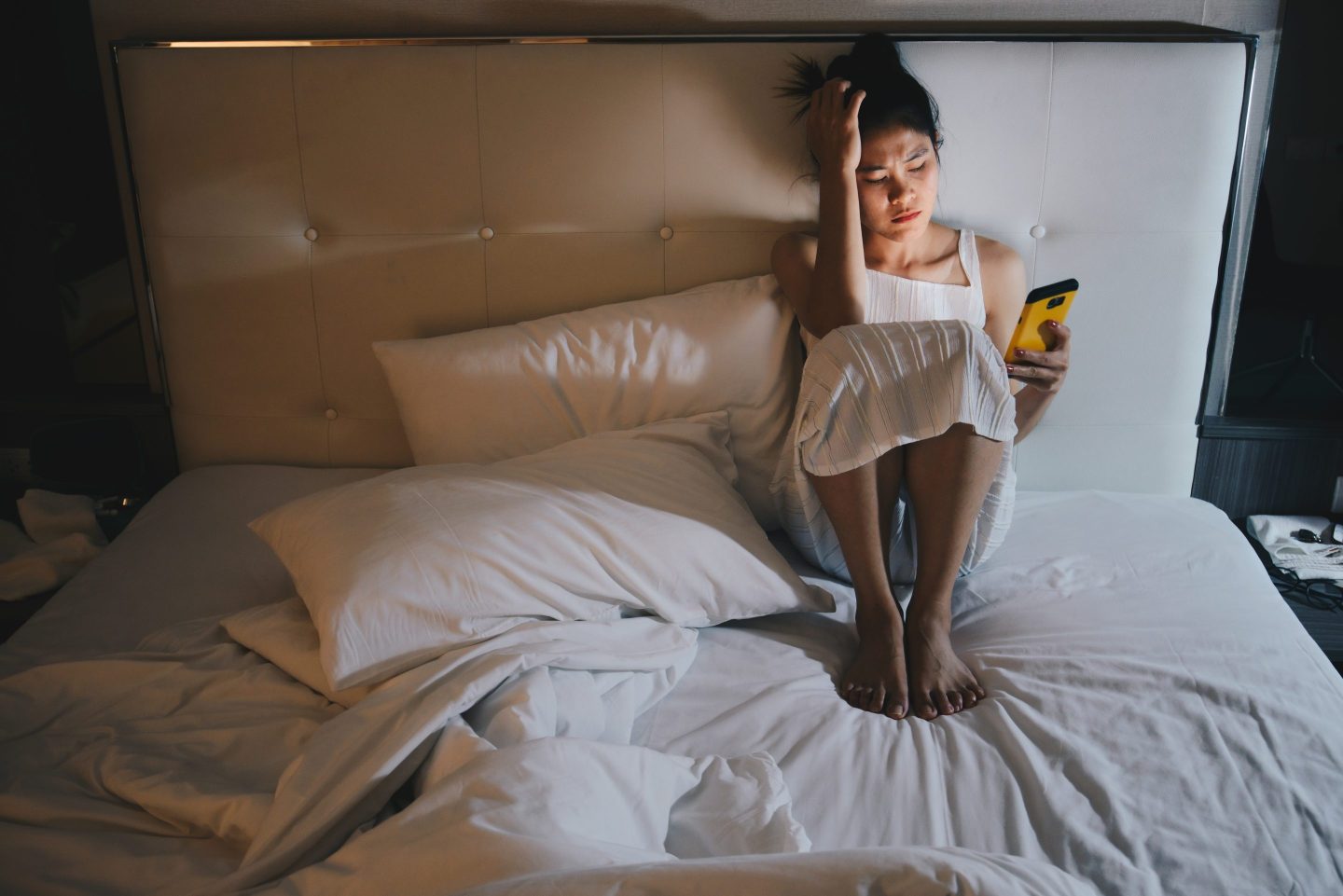 Woman sitting alone on the bed in the bedroom and looking to mobile phone in her hand.
